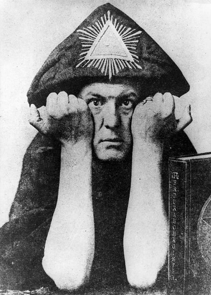 Black and white photo of Aleister Crowley wearing a ceremonial robe and headdress with a radiant triangle symbol. He rests his head on his fists and stares directly at the camera.