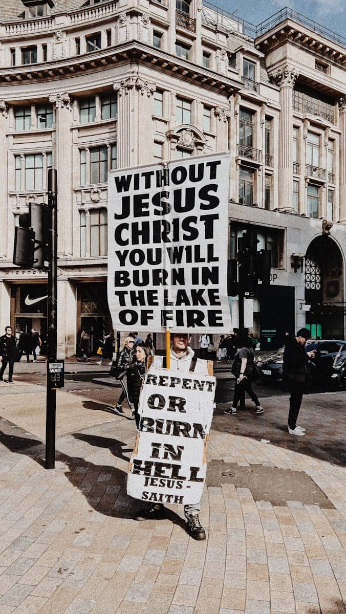 Street preacher in a busy city centre holding large signs that read “Without Jesus Christ you will burn in the lake of fire” and “Repent or burn in hell.”