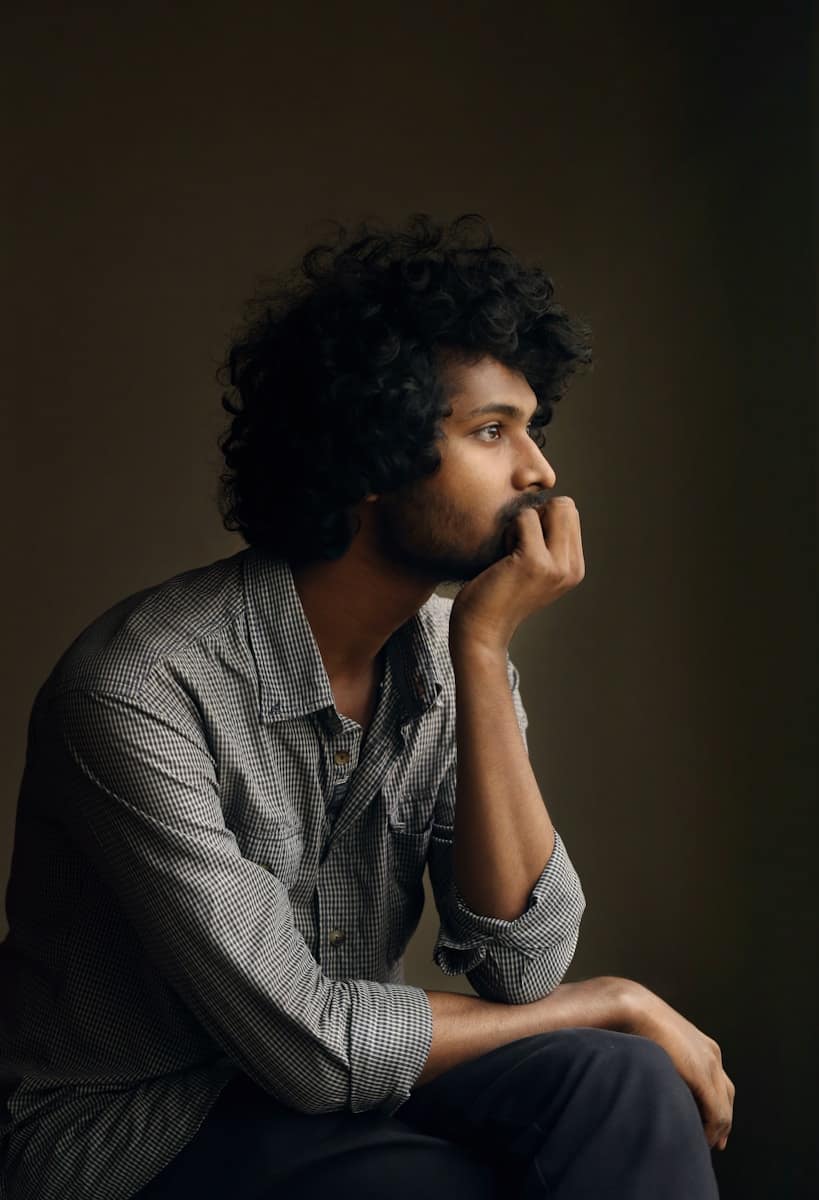 man in gray and white checkered dress shirt, A thoughtful person sitting with their hand on their chin, deep in reflection, illustrating what does tarot help with during times of uncertainty.