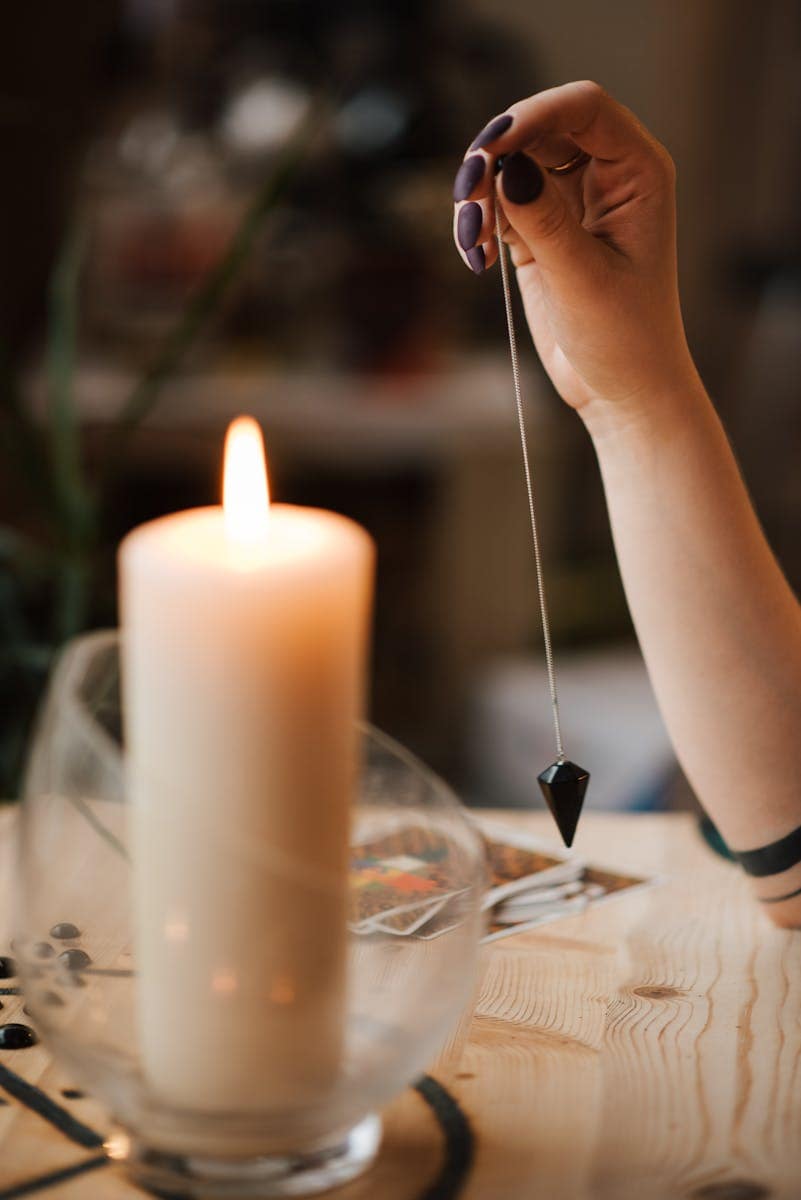 A hand holds a pendulum over tarot cards by candlelight, evoking a mystical ambiance.