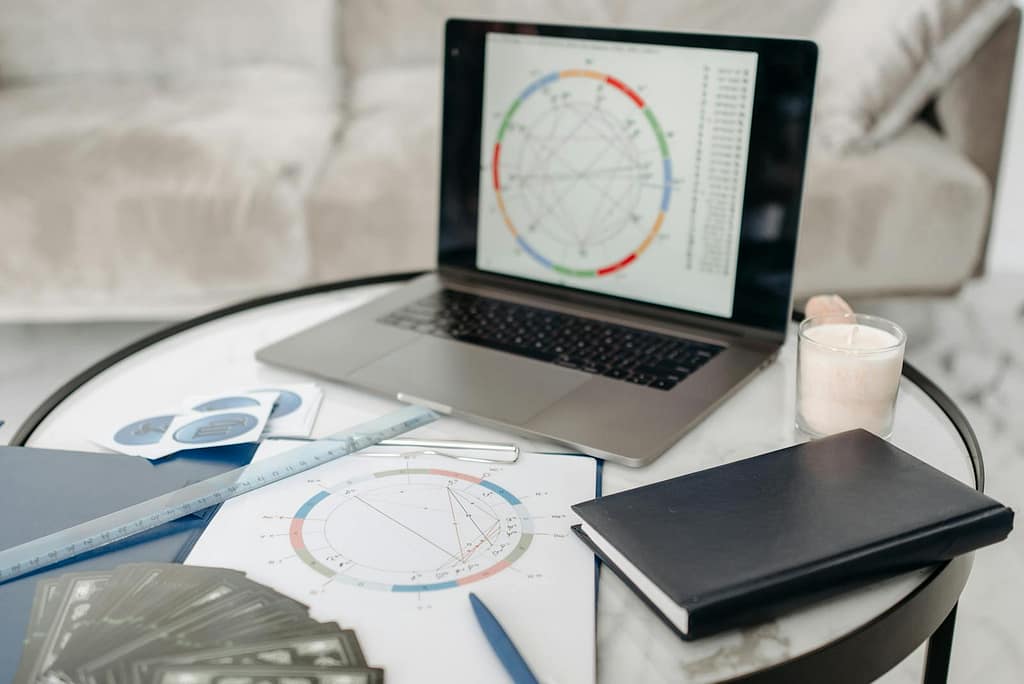 Astrology charts displayed on a laptop next to tarot cards and notebooks on a table.