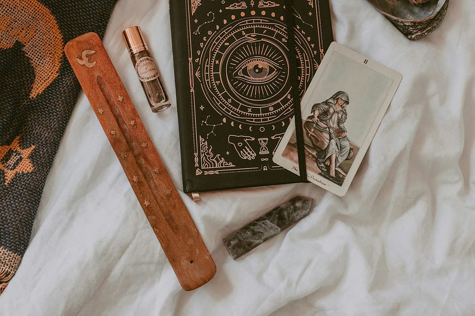 Flat lay of spiritual tools including a tarot card, black journal with mystical design, incense holder, sage bundle, and crystals, representing tarot, journalling and meditation in Manchester.